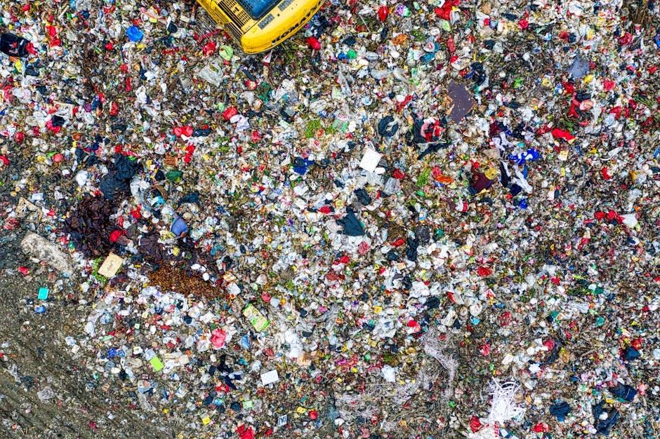 An extensive aerial view of a large landfill site filled with a dense accumulation of mixed waste materials. The waste consists primarily of plastics, including bottles, bags, and fragments of various